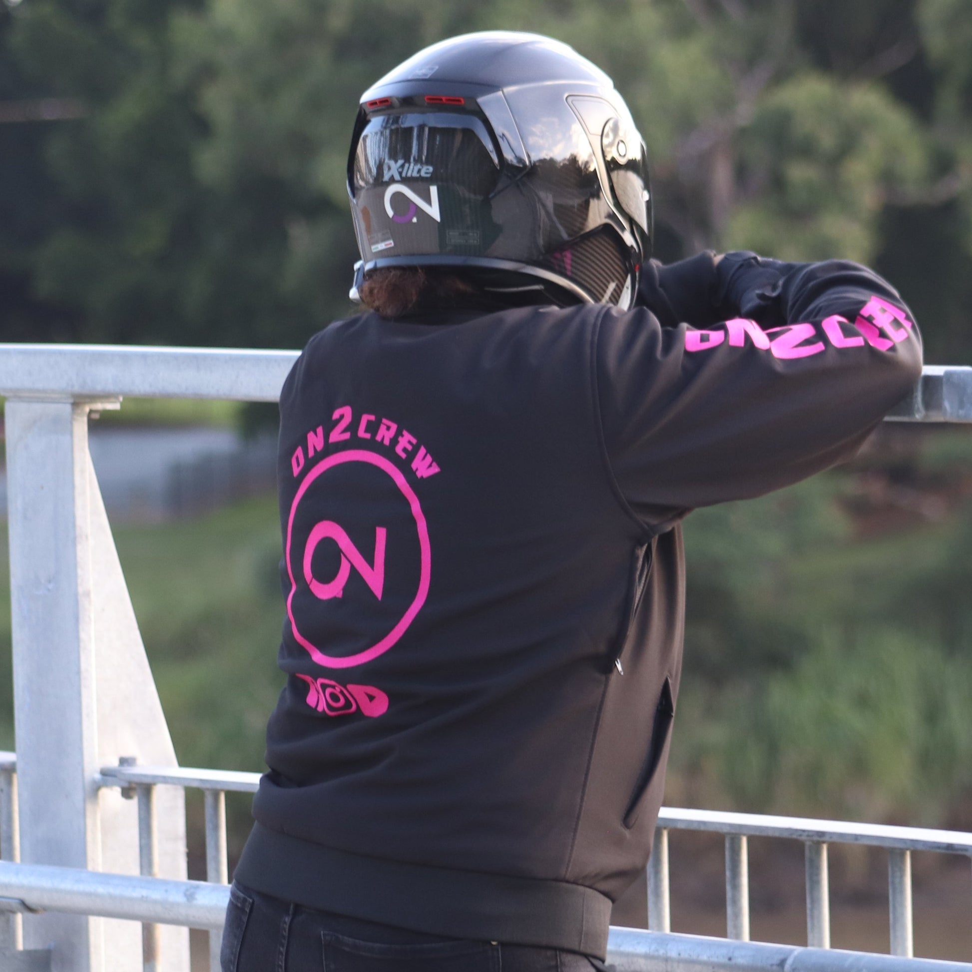 A person wearing a pink motorcycle riding hoodie with the On2Crew logo, riding a motorcycle.