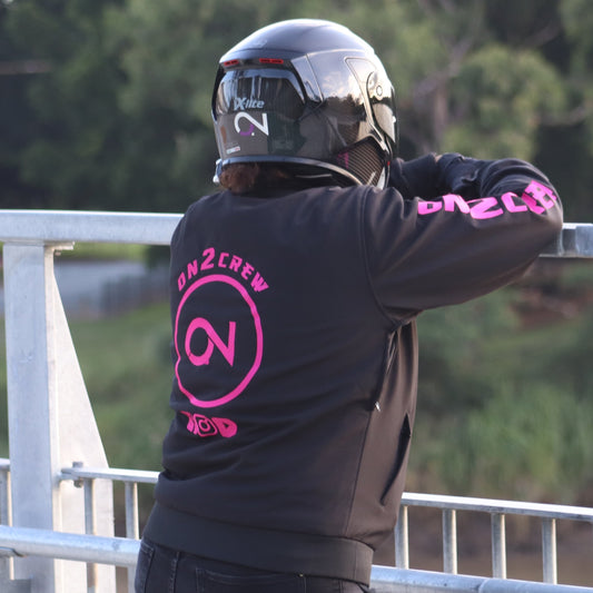 A person wearing a pink motorcycle riding hoodie with the On2Crew logo, riding a motorcycle.